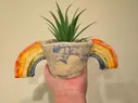 A fired purple plant pot with rainbow arms coming from either side, with a fake plant in it