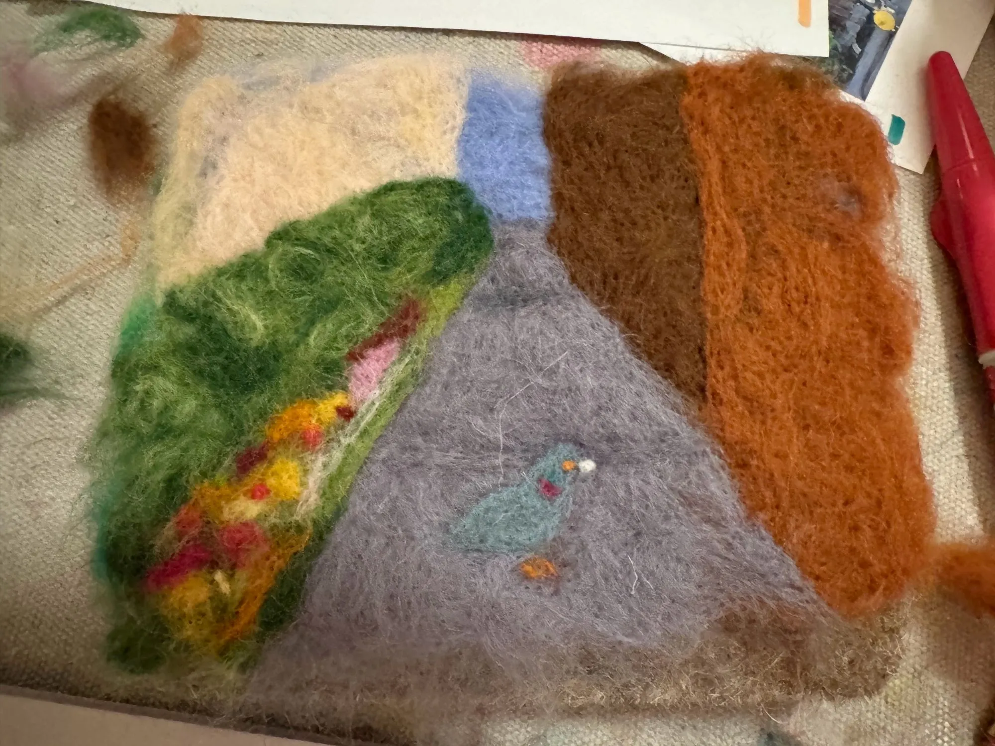 A needle-felted scene showing a pigeon on a city sidewalk next to some flowers and bushes