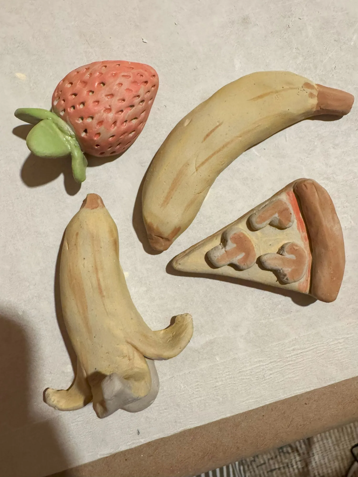Four unfired clay pieces shaped like a peeled banana, an unpeeled banana, a strawberry, and a slice of mushroom pizza
