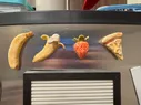 Four clay magnets on a fridge, shaped like a peeled banana, an unpeeled banana, a strawberry, and a slice of mushroom pizza
