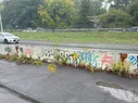 A barrier between a path and road with various graffiti on a rainy day