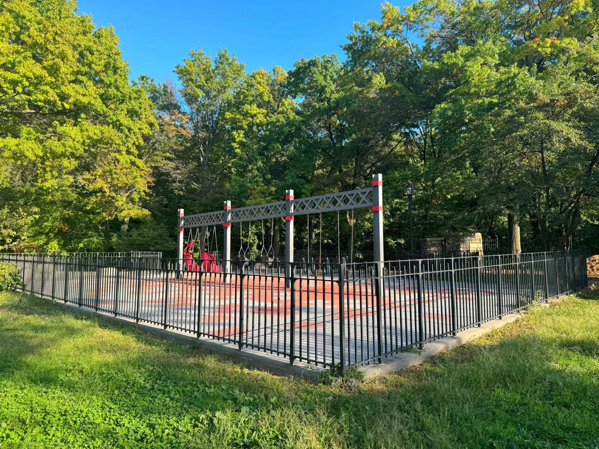 A swingset in a fenced playground surrounded by green trees