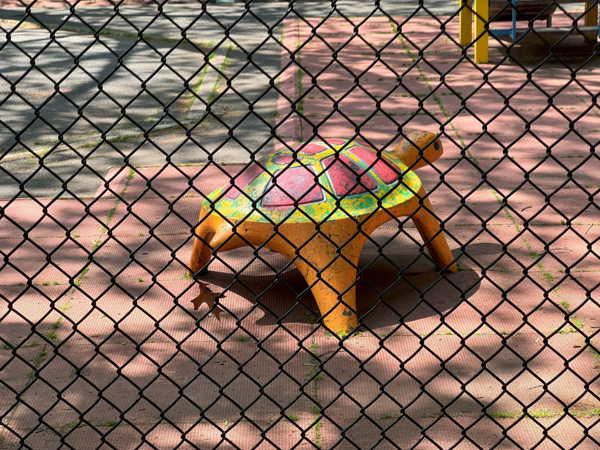 A turtle structure with faded paint on a playground, visible through a chain-link fence