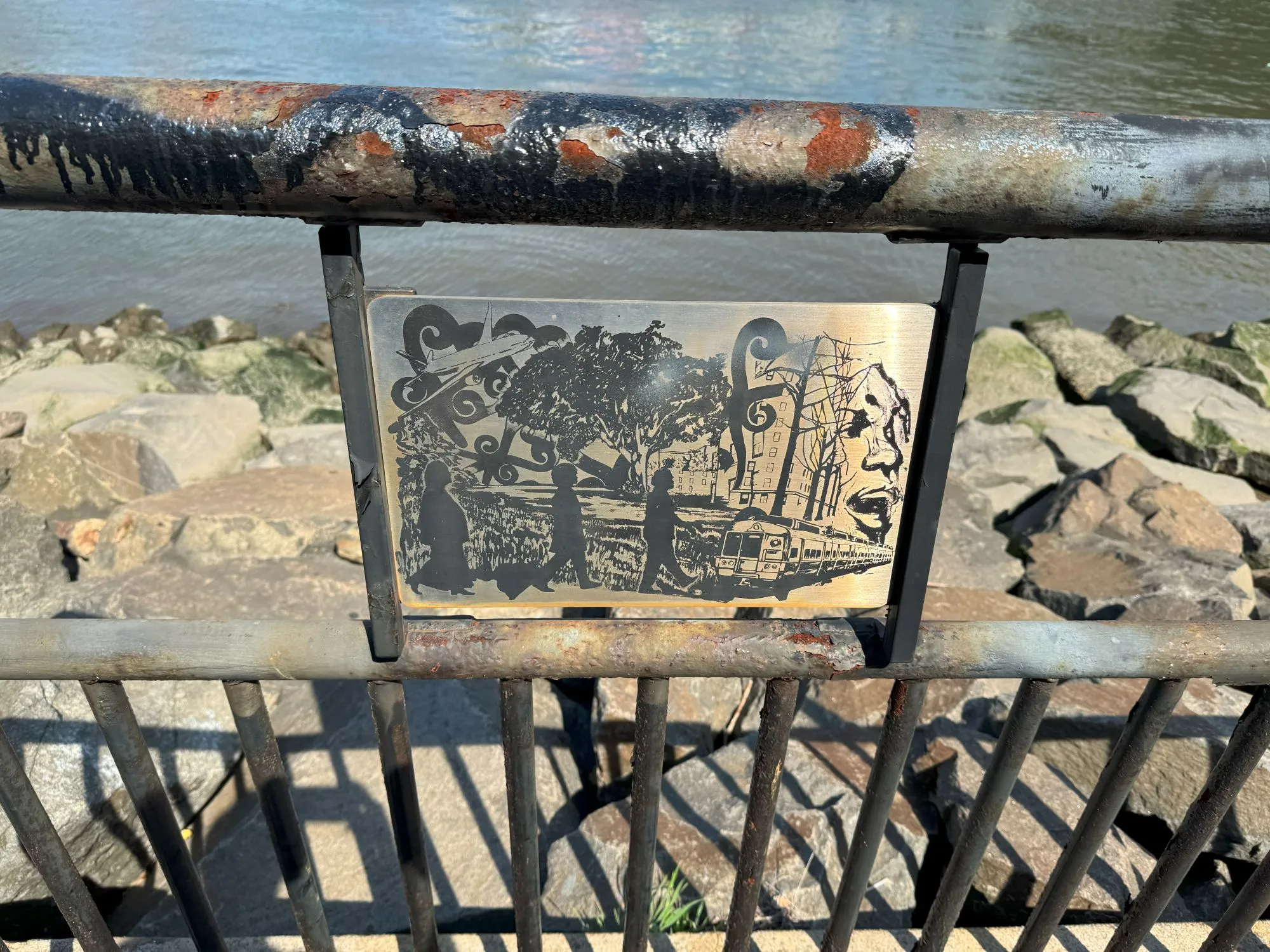 A monochrome art piece on metal, mounted on a fence next to the river