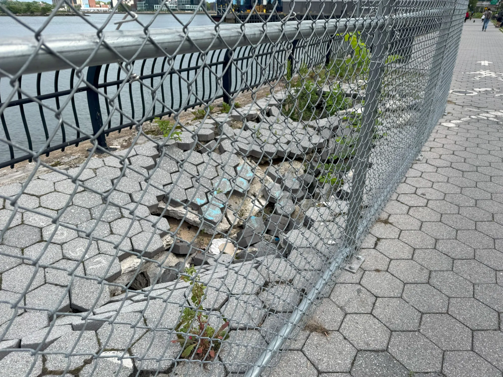 A broken section of path along the river, behind a chain-link fence