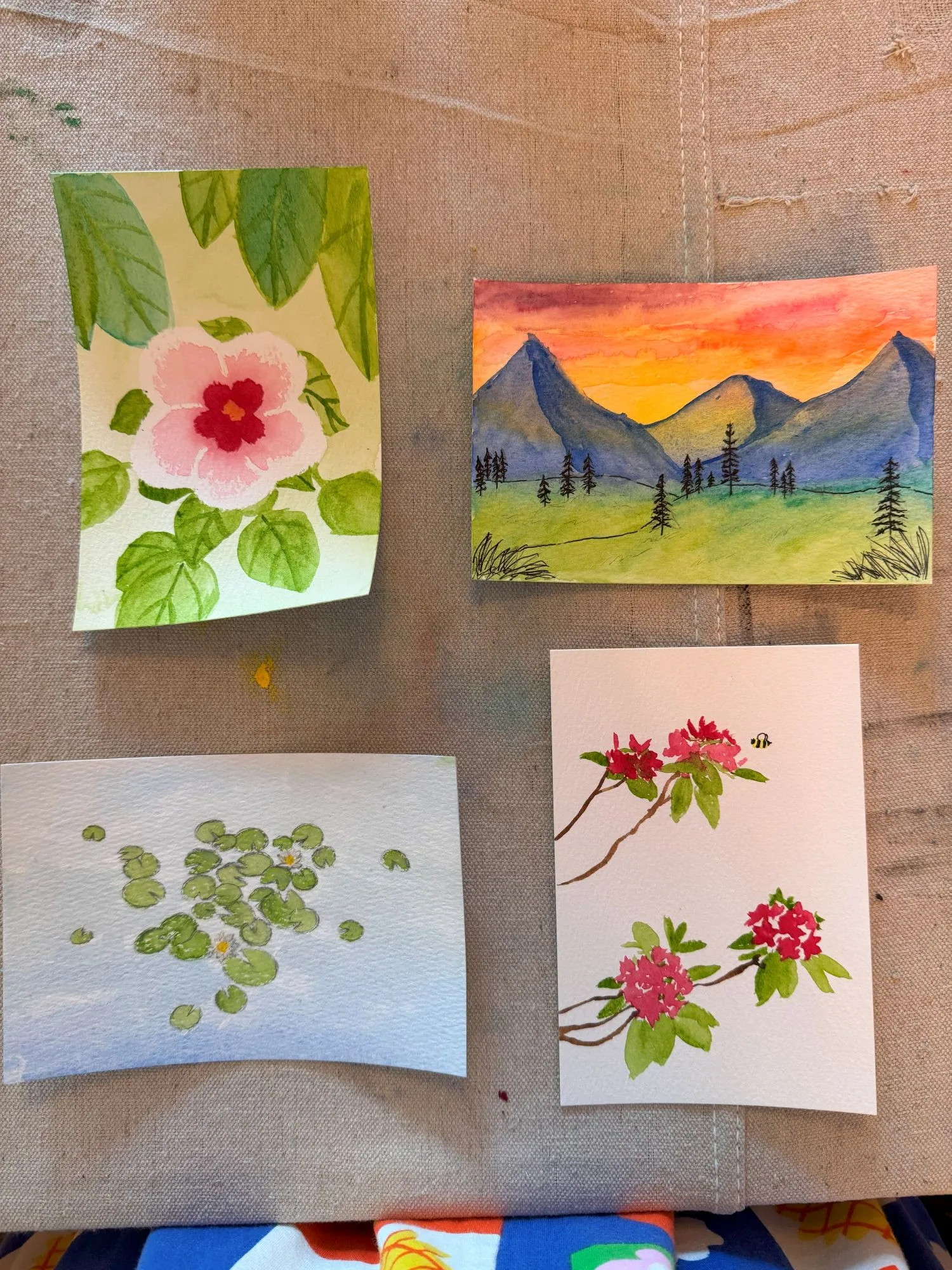 Four small watercolor paintings. One is of a mountainous landscape, one is of a flower, one is of flowers and a bee, and one is of lily pads